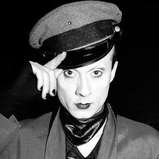 Klaus Nomi Black and White image of the singer Klaus Nomi. He is wearing black lipstick while looking to the camera with a blank expression on his face. He wears black gloves, a military-like hat, and a black bandana.