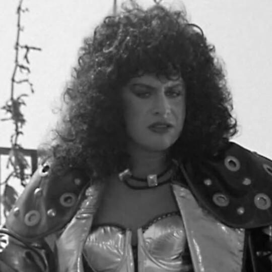 A Black and White image of the character Velvet Von Ragnar from the movie Never Too Young to Die with a serious expression. She has black curly hair, eyeshadow, lipstick on, a necklace and corset, and a black leather jacket