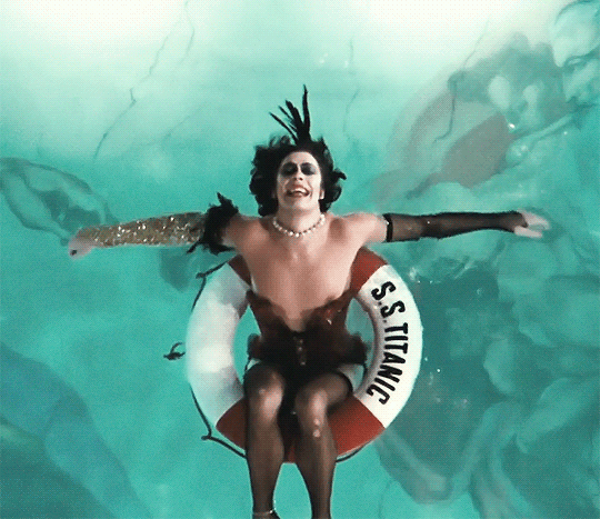 Gif of the character Dr Frank-N-Furter from the movie Rocky Horror Picture Show. He is swimming in a pool with teal water while sitting in a buoy that has "S.S. TITANIC" on the side. His makeup is running down his face while he sings.